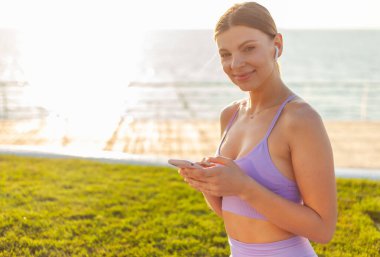 Sabah sporu. Spor giyim sektöründe genç bir bayan gün doğumunda sahilde otururken akıllı telefon kullanıyor.