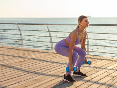Sabah sporu. Spor kıyafetli genç, sıska bir kadın gün doğumunda elinde dambıllarla plajda çömelme egzersizi yapıyor.