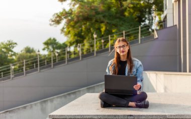 Merdivenlerde dizüstü bilgisayarla oturan kot ceketli, modern bir bayan öğrenci. Mesafe öğrenme. Modern gençlik kavramı.