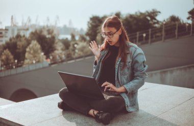 Kot ceketli bir kadın öğrenci şehirde açık havada merdivenlerde otururken video izliyor..