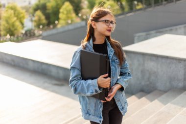 Kot ceketli ve gözlüklü genç bayan şehirde elinde dizüstü bilgisayarla merdivenlerden çıkıyor.