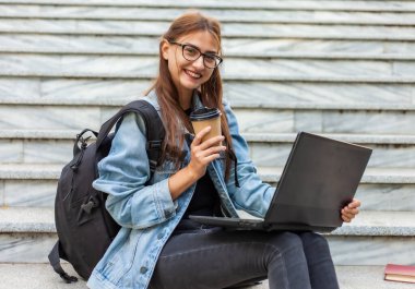 Kot ceketli ve sırt çantalı mutlu modern bayan öğrenci dışarıda dizüstü bilgisayarla merdivenlerde oturuyor. Kahve içiyormuş. Mesafe öğrenme. Modern gençlik kavramı.