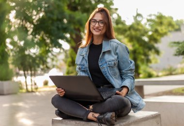 Merdivenlerde dizüstü bilgisayarla oturan kot ceketli, modern bir bayan öğrenci. Mesafe öğrenme. Modern gençlik kavramı.