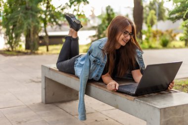 Çevrimiçi çağrı. Mesafe öğrenme. Kot ceketli ve gözlüklü genç bir bayan öğrenci parktaki bankta yatarken dizüstü bilgisayara bakıyor.