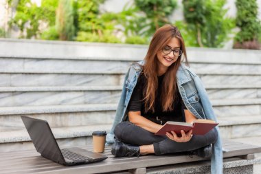 Modern öğrenciler. Mesafe öğrenme. Genç hevesli kadın dizüstü bilgisayarla bankta otururken kitap okuyor.