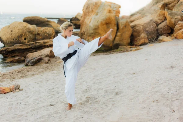 Young attractive karate woman in white kimono with a black belt trains kicks on a wild beach with stones