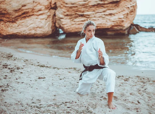 Attractive female martial artist in a white kimono with a black belt stands in fighting stance on wild beach