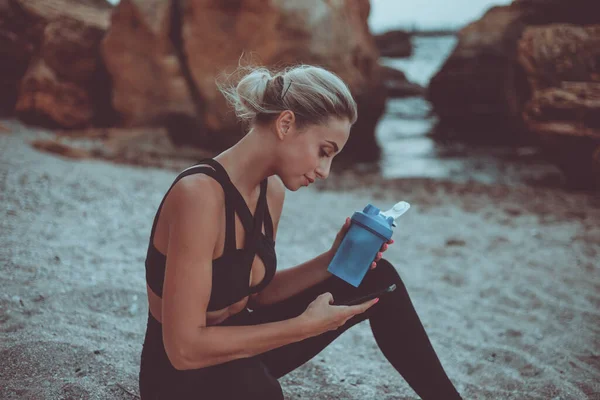 Young attractive fit woman in sportswear is resting after training while sitting on the sand at the wild beach. Sports woman uses a smartphone and drinks water from a bottle