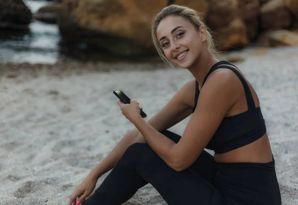 若いですフィットブロンド女性でスポーツウェア座っていますザ砂の上に野生のビーチとホールドスマートフォンで彼女の手 — ストック写真