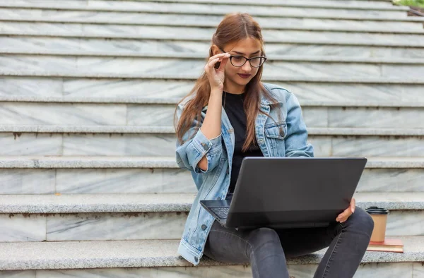 Young modern woman student in a denim jacket sitting on the stairs with laptop. Watching video. Distance learning. Modern youth concept.