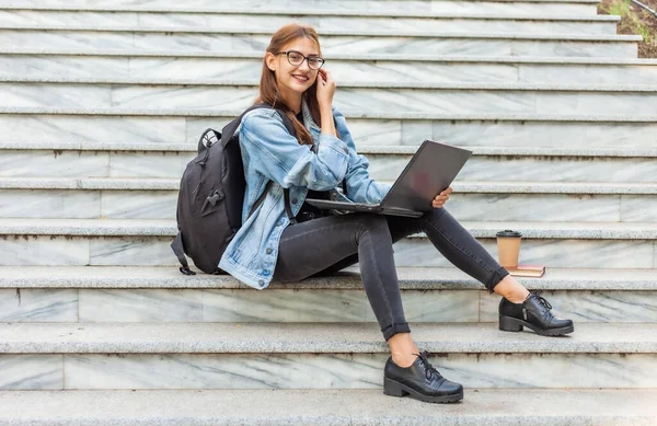 Happy modern woman student in denim jacket and backpack sitting on the stairs with laptop outdoor. Distance learning. Modern youth concept.