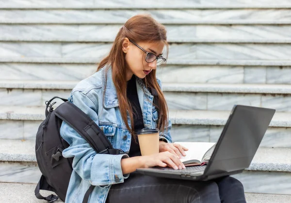 Happy modern woman student in denim jacket and backpack sitting on the stairs and uses laptop with book outdoor. Distance learning. Modern youth concept.