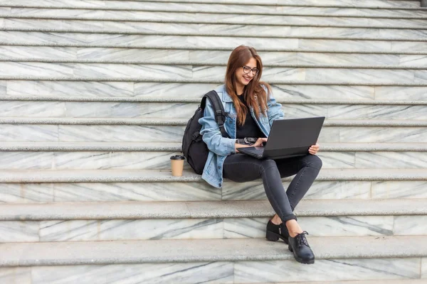 Young modern woman student in a denim jacket sitting on the stairs with laptop. Distance learning. Modern youth concept.
