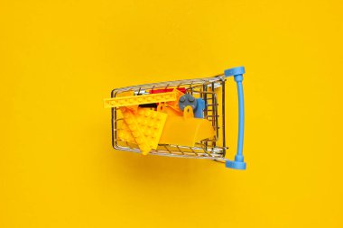 Sarı arka planda oyuncak tuğlaları olan küçük bir alışveriş arabası. Çocuk alışverişi konsepti. Üst görünüm