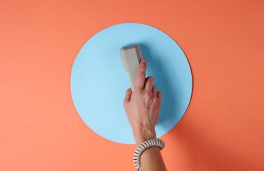 Kabloya sarılı kadın el, mavi daireli mercan rengi arka plana karşı telefon tüpü tutuyor. Liman sanatı. Yaratıcı minimalizm. Üst görünüm