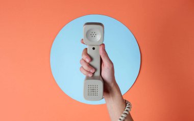 Kabloya sarılı kadın el, mavi daireli mercan rengi arka plana karşı telefon tüpü tutuyor. Liman sanatı. Yaratıcı minimalizm. Üst görünüm