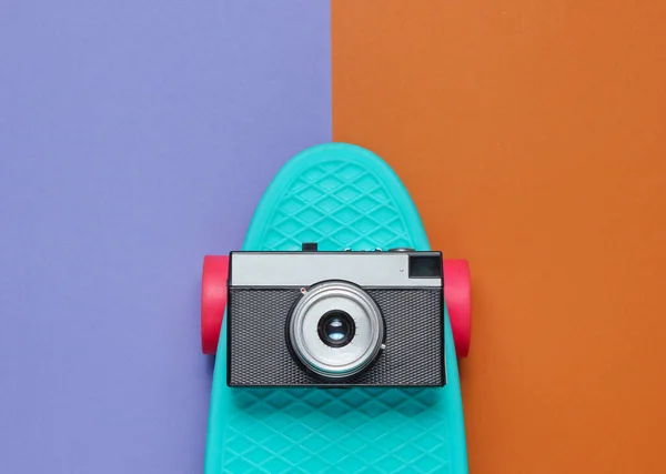 Pop art retro style still life. 80s. Cruiser board with retro camera on two-color paper background. Summer fun. Top view.