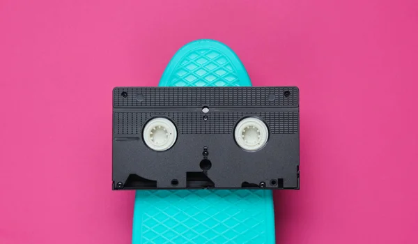 Pop art retro style still life. 80s. Cruiser board and video cassette on pink paper background. Summer fun. Top view.