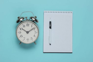 Mavi arka planda çalar saati olan bir defter. Minimalist iş konsepti. Üst görünüm