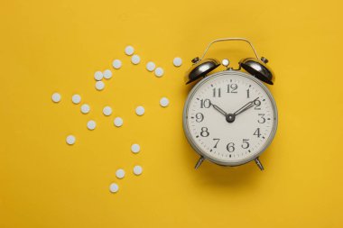 İlaç alma zamanı. Sarı arka planda haplar ve çalar saat. Üst görünüm