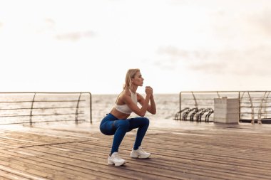 Deniz kenarındaki güvertede sağlıklı kadın eğitimi. Spor giyimli kadın gün doğumunda plajda spor lastiğiyle çömelme egzersizi yapıyor.