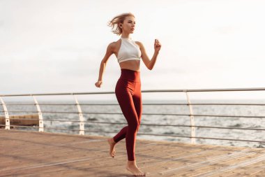Sabah koşusu. Spor giyimli genç bir kadın güneş doğarken plajda koşuyor. Açık hava antrenmanı. Sağlıklı yaşam tarzı