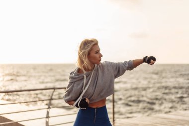 Sargı bezine sarılmış elleri olan genç bir kickboksör kadın gün doğumunda sahilde trenleri yumrukluyor. Sabah dövüşçüsü