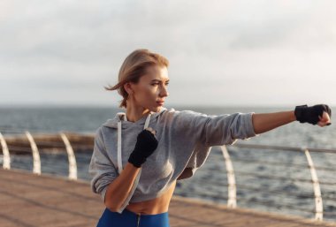 Sargı bezine sarılmış elleri olan genç bir kickboksör kadın gün doğumunda sahilde trenleri yumrukluyor. Sabah dövüşçüsü