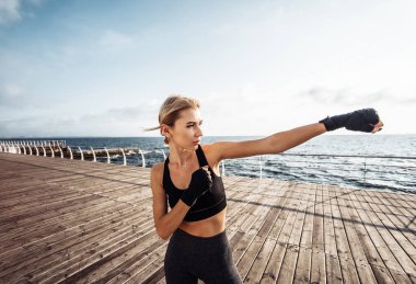 Sargı bezine sarılmış elleri olan genç bir kickboksör kadın gün doğumunda sahilde trenleri yumrukluyor. Sabah dövüşçüsü