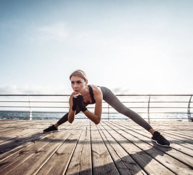 Deniz kenarındaki güvertede sağlıklı kadın eğitimi. Spor giyim sektöründe genç ve çekici bir kadın güneş doğarken plajda egzersiz yapmadan önce esneme hareketleri yapar. Isının.