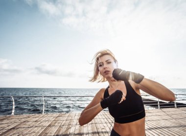 Sargı bezine sarılmış elleri olan genç bir kickboksör kadın gün doğumunda sahilde trenleri yumrukluyor. Sabah dövüşçüsü