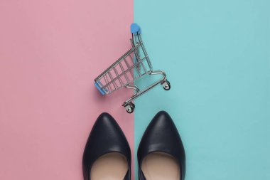 Minimalist moda ve alışveriş konsepti. Deri yüksek topuklu ayakkabılar, mavi-pembe pastel arka planda alışveriş arabası. Üst görünüm