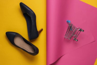 Minimalist moda ve alışveriş konsepti. Deri yüksek topuklu ayakkabılar, sarı pastel arka planda alışveriş arabası. Üst görünüm