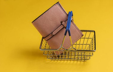 Minimalist alışveriş konsepti. Sarı arka planda mini alışveriş sepeti ve deri cüzdan..