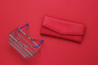 Minimalist alışveriş konsepti. Kırmızı arka planda mini alışveriş sepeti ve kırmızı deri cüzdan. Üst görünüm