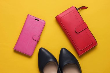 Stüdyo moda çekimi. Yüksek topuklu ayakkabılar, kırmızı cüzdan, sarı arka planda deri çantalı akıllı telefon. Üst görünüm