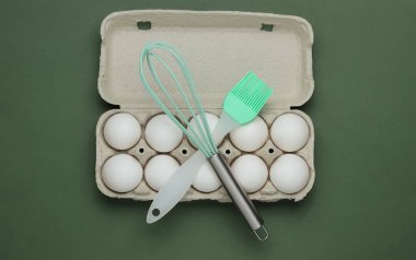 Yemek pişirme kavramı. Yeşil arka planda karton yumurta tepsisi, mutfak aletleri (çırpma, fırça). Üst görünüm