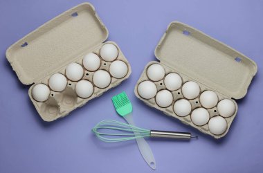 Yemek pişirme kavramı. Mor arka planda karton yumurta tepsisi, mutfak aletleri (fırça, fırça). Üst görünüm