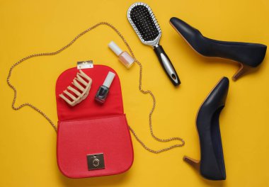 Kadın çantasında ne var? Kadın aksesuarlarıyla kırmızı deri çanta, kozmetik ürünleri, sarı arka planda yüksek topuklu ayakkabılar. Minimalist moda konsepti. Stüdyo çekimi
