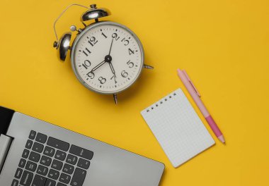 Laptop, çalar saat, sarı arka planda not defteri olan pembe kalem. Üst görünüm