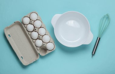 Yemek pişirme konsepti. Boş beyaz kase çırpılmış, yumurta tepsisi mavi stüdyo arka planında. Üst görünüm.