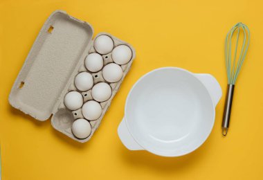Yemek pişirme konsepti. Boş beyaz kase çırpılmış, sarı stüdyo arka planında yumurta tepsisi. Üst görünüm.