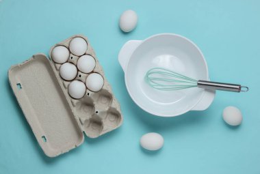 Yemek pişirme konsepti. Boş beyaz kase çırpılmış, yumurta tepsisi mavi stüdyo arka planında. Üst görünüm.