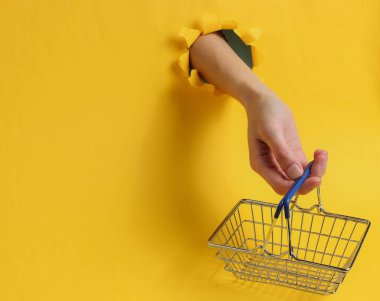 Minimalizm alışveriş konsepti. Kadın el, sarı arka plandaki yırtık bir delikten alışveriş sepeti tutuyor.