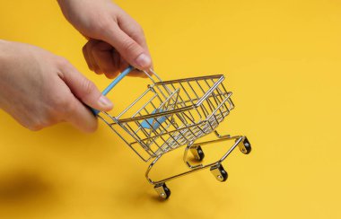 Minimalizm alışveriş konsepti. Kadın elleri sarı arka planda mini alışveriş arabası tutuyor.