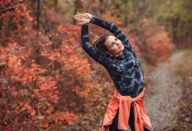 Spor giyimli genç ve çekici bir kadın sonbahar ormanında ağaçların kızıl yapraklarıyla el egzersizi yapıyor. Eğitimden önce ısın. Sağlıklı yaşam tarzı kavramı