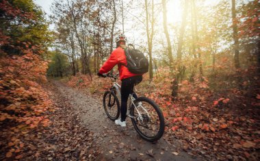 Tanınmayan bisiklet bisikletçisi kadın, orman yolunda ağaçların kızıl yapraklarıyla geziyor..