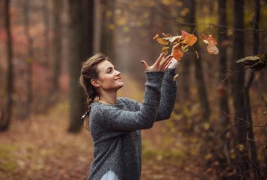 Örgülü örgülü örgülü saçları olan mutlu genç bir kadın sonbahar ormanında eline düşen yaprakları fırlatıyor. Sonbahar sanat portresi