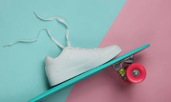 Stylish white sneakers with untied laces and cruiser board on  pink blue pastel background. Summertime fun. Studio shot, top view.