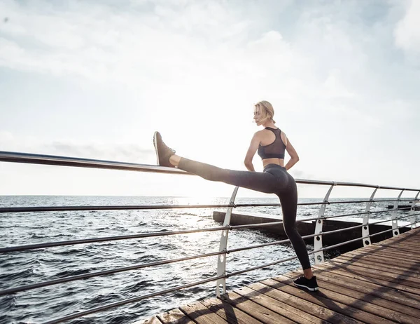Deniz kenarındaki güvertede sağlıklı kadın eğitimi. Spor giyim sektöründe genç ve çekici bir kadın gün doğumunda plajda egzersiz yapmadan önce bacak esnetiyor. Isının.